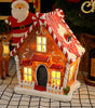 Decorative gingerbread house with lit windows on a checkered surface, surrounded by Christmas-themed items.