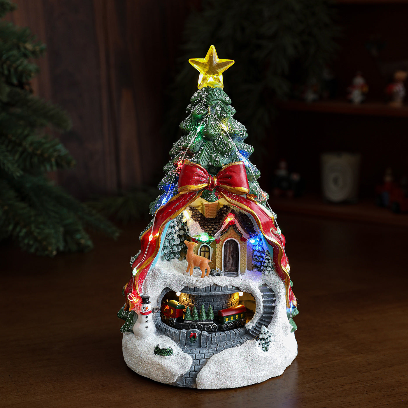 Decorative Christmas scene with a lit-up tree, house, and reindeer on a wooden surface.
