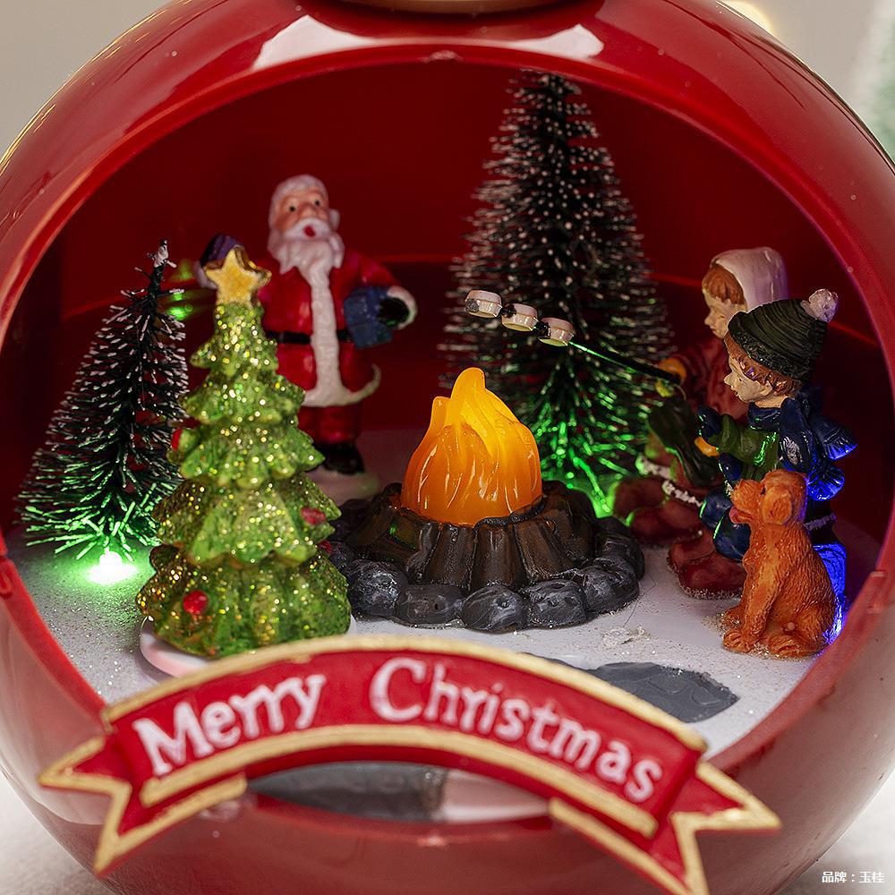 Christmas-themed ornament with Santa Claus, trees, and a family around a campfire, featuring a 'Merry Christmas' banner.