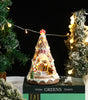 Decorative Christmas tree-shaped gingerbread house with lights, surrounded by festive trees and decorations.